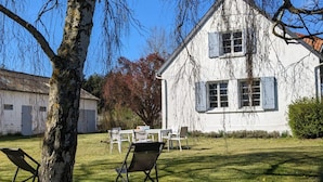 A white building with a sloped roof and shutters, a lawn with chairs, and a tree in the foreground. - Anna & Jules' little house (Saint-Quentin-en-Tourmont)