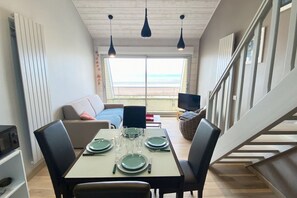 Ein Essbereich mit einem Tisch für vier Personen, einer Couch, einem Fernseher und Blick auf den Strand.