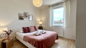 Une chambre à coucher avec un lit, des tables de chevet, une lampe, une fenêtre avec des rideaux et des œuvres d’art accrochées au mur.