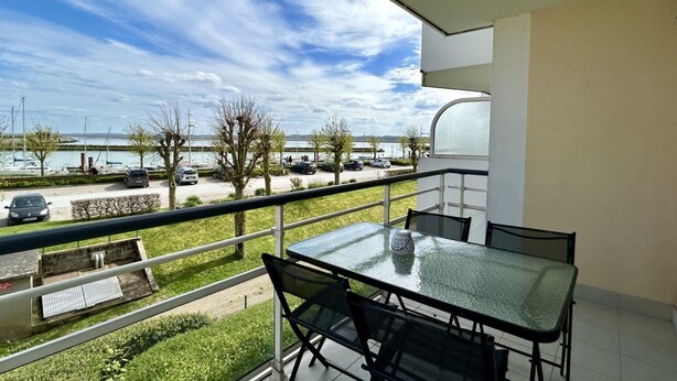 Un balcon avec une table et des chaises en verre, donnant sur un parking et une étendue d’eau.