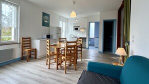A modern kitchen with white cabinets, a dining table with wooden chairs, and a blue sofa in the living area. - La pommeraie 4, apartment for 4 people (Quend)