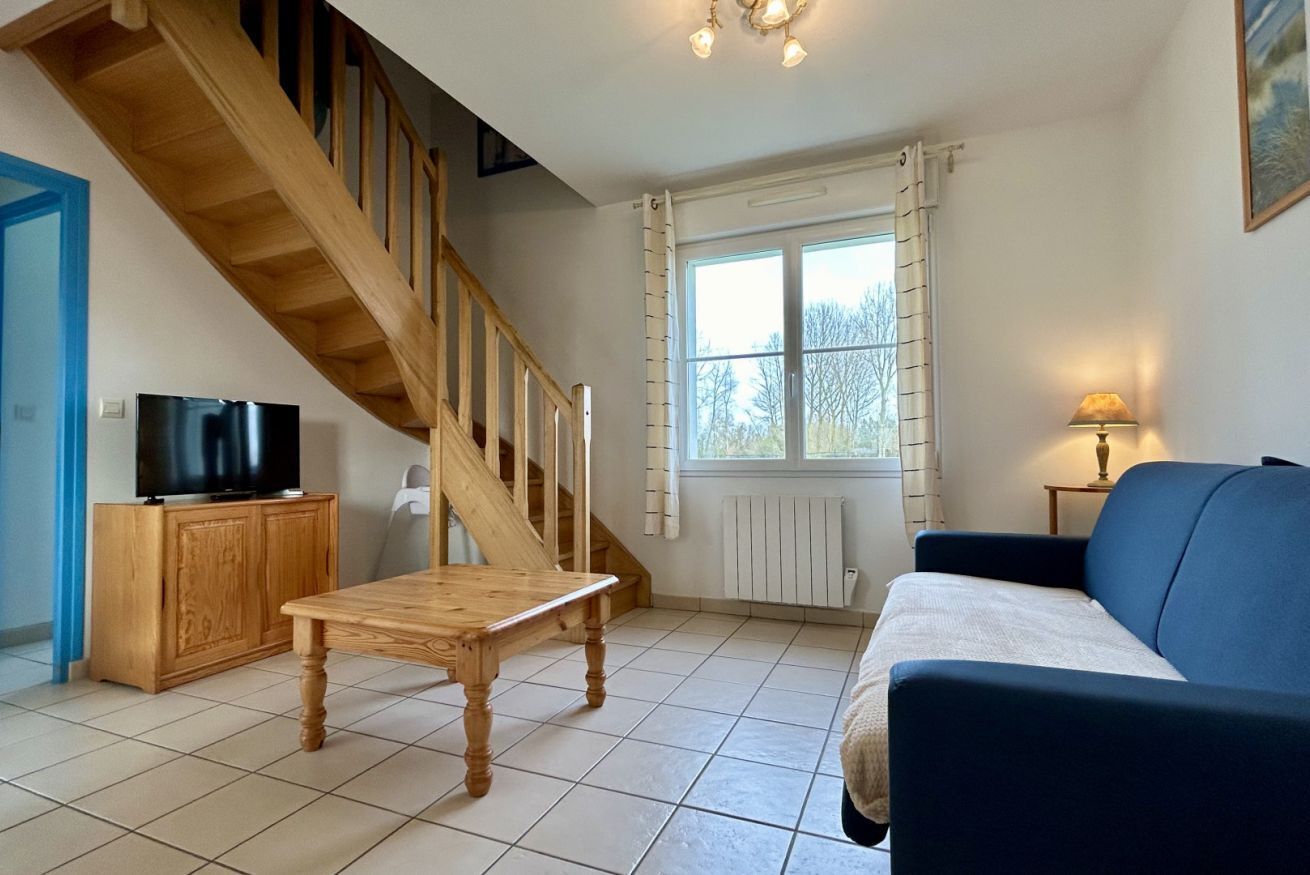 Un salon avec un escalier en bois, un téléviseur posé sur un meuble, un canapé et une fenêtre avec des rideaux.