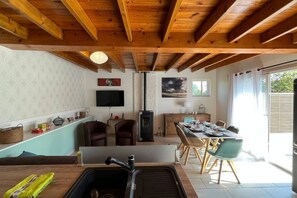A kitchen with wooden beams, a TV, a fireplace, a dining area with chairs, and a window.