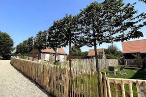 Un camino de grava conduce a una cerca de madera con un portón de metal, un área verde con una silla y un edificio de techo rojo al fondo.