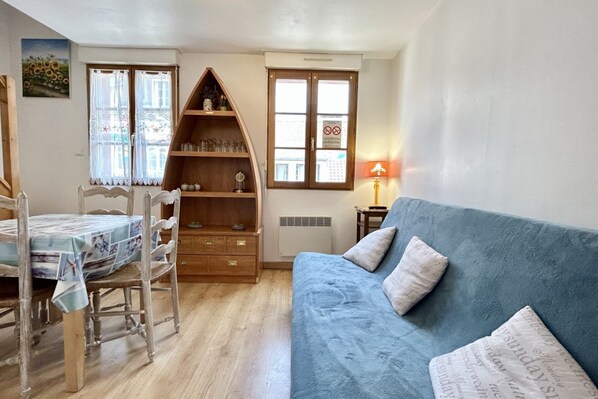 A room with a dining table, chairs, a blue sofa, a bookshelf, and a window with a view. - Gîte 4, 3-person gîte in the center of town (Le Crotoy)