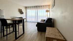Een moderne woonkamer met een bank, een eettafel met stoelen, een keuken met een koelkast, en een groot raam met dunne gordijnen.