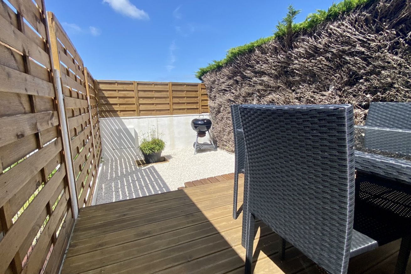 Eine Holzterrasse mit einem Korbstuhl, einem Grill und einem kleinen Blumentopf.
