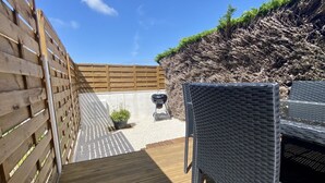 Una terraza de madera con una silla de mimbre, una parrilla y una pequeña planta en maceta.