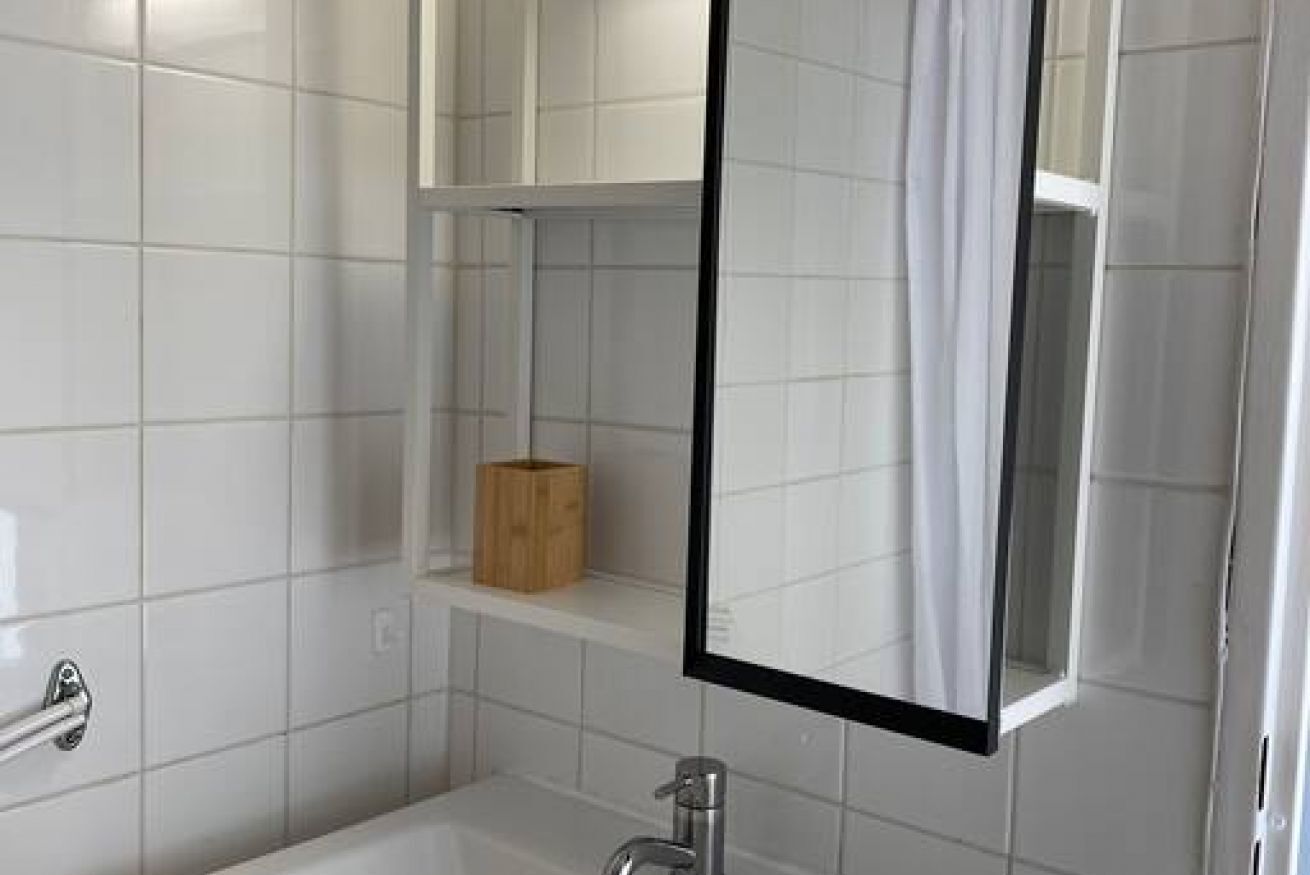 A bathroom with white tiles, a bathtub, a mirror, and a wooden shelf.