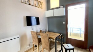Una habitación con una mesa de comedor de madera, dos sillas, un televisor montado en la pared, un armario blanco y una puerta que da a un área exterior.