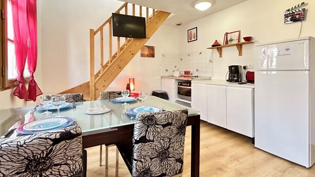 Un coin repas avec une table en verre, des chaises à motifs floraux, un escalier en bois, une cuisine avec des meubles blancs, un réfrigérateur blanc et un téléviseur fixé au mur.