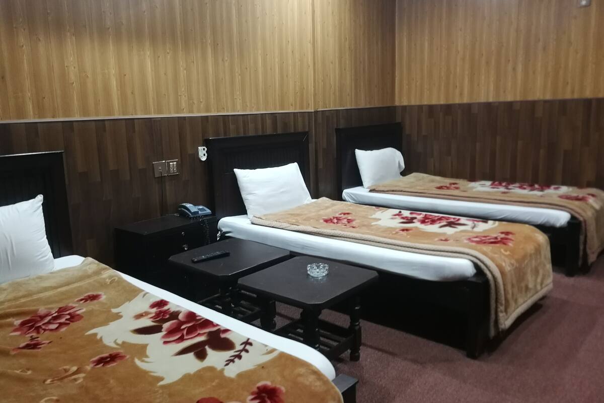 Ein Hotelzimmer mit drei Betten, jeweils mit einem blumigen Überwurf, einem Nachttisch mit Fernbedienung und einem kleinen Tisch mit einem Glas.