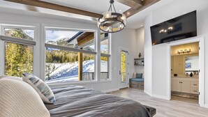 Ett sovrum med en stor säng, en tv som är monterad på väggen och utsikt över ett snötäckt berglandskap genom stora fönster.