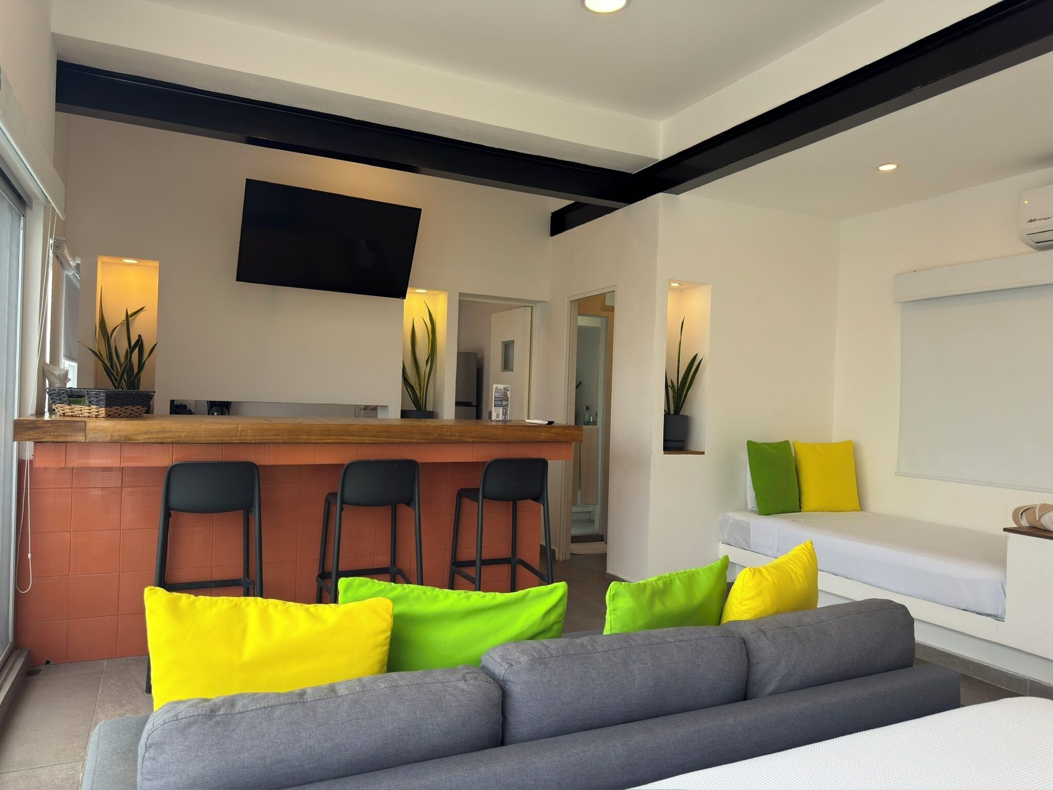 A modern hotel room with a grey sofa, a wooden bar area with bar stools, a flat-screen TV mounted on the wall, and a bed with a white mattress and grey headboard.