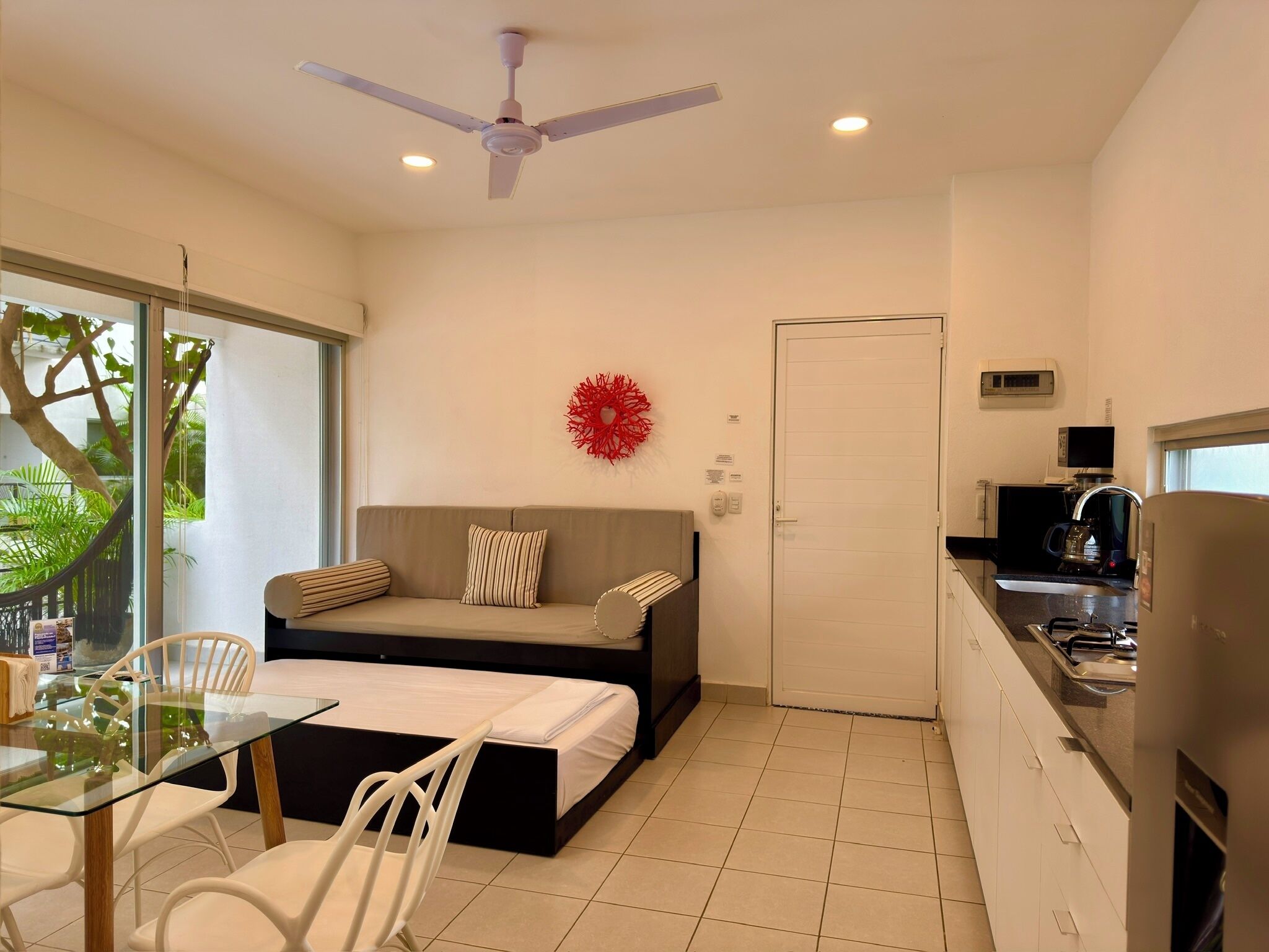A compact hotel room with a bed, a sofa, a dining table with chairs, a kitchen area, and a ceiling fan.