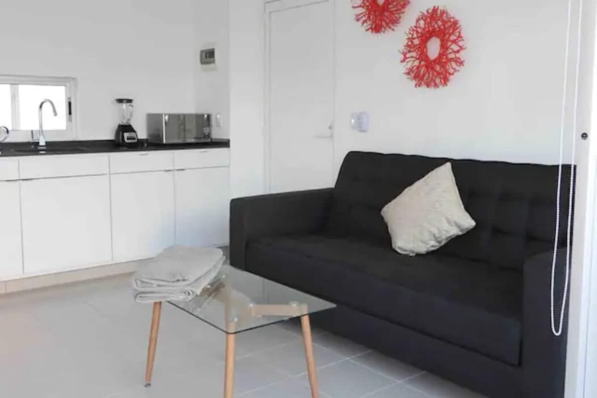 Una sala moderna con un sofá negro, una mesa de centro de vidrio y una cocina con armarios blancos y decoración mural roja.