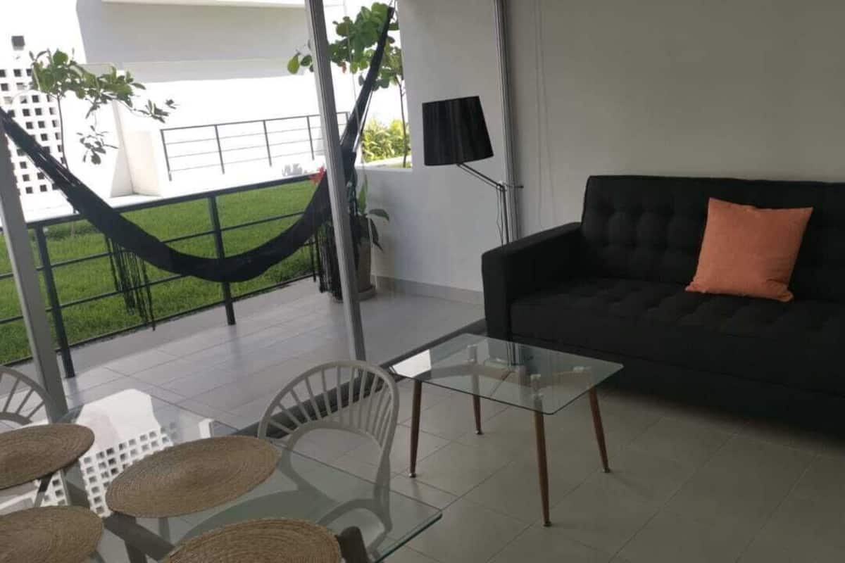Una sala de estar con una mesa de centro de vidrio, un sofá oscuro con un cojín, una hamaca y vistas a un edificio blanco y vegetación.