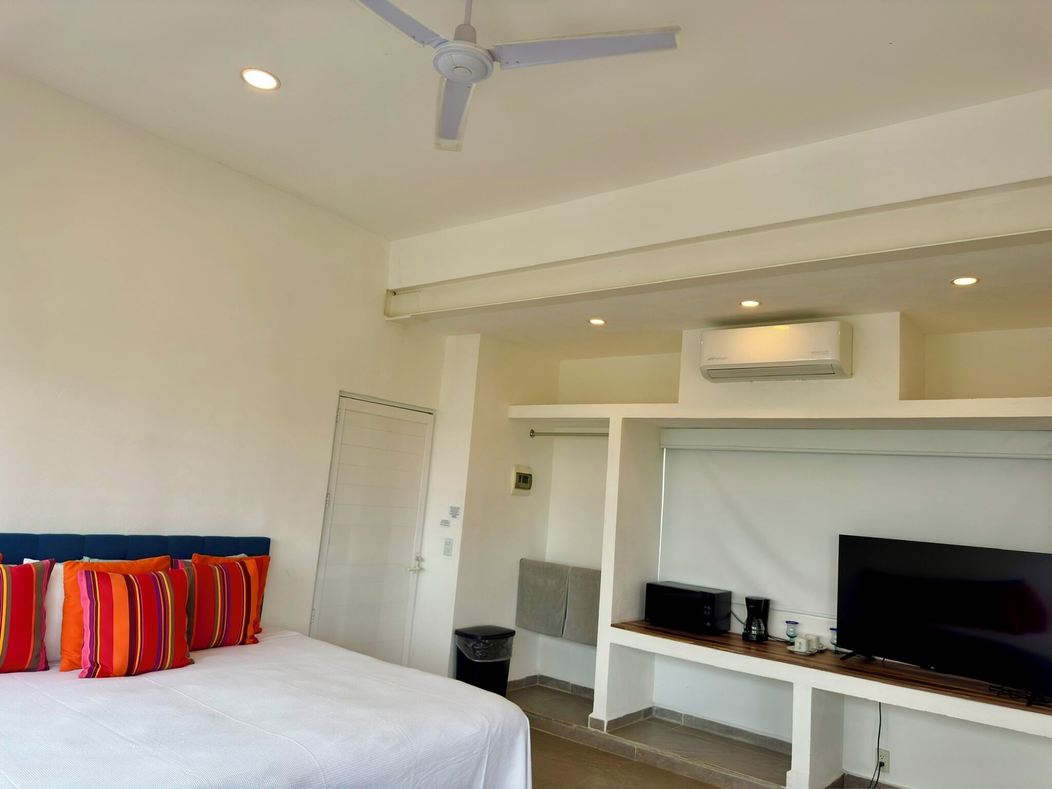 Una habitación de hotel con una cama, almohadas coloridas, un ventilador de techo, un equipo de aire acondicionado, un televisor sobre una estantería de madera y un cesto de basura.