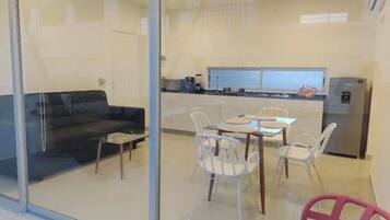 Een modern interieur met een glazen scheidingswand tussen een zithoek en een keuken. Er is een eettafel met stoelen, een bank en een koelkast.