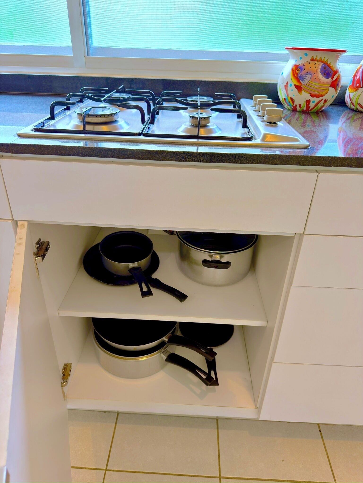 Une cuisine équipée d’une cuisinière à gaz, de meubles blancs et de deux étagères où sont rangées des casseroles et des poêles.