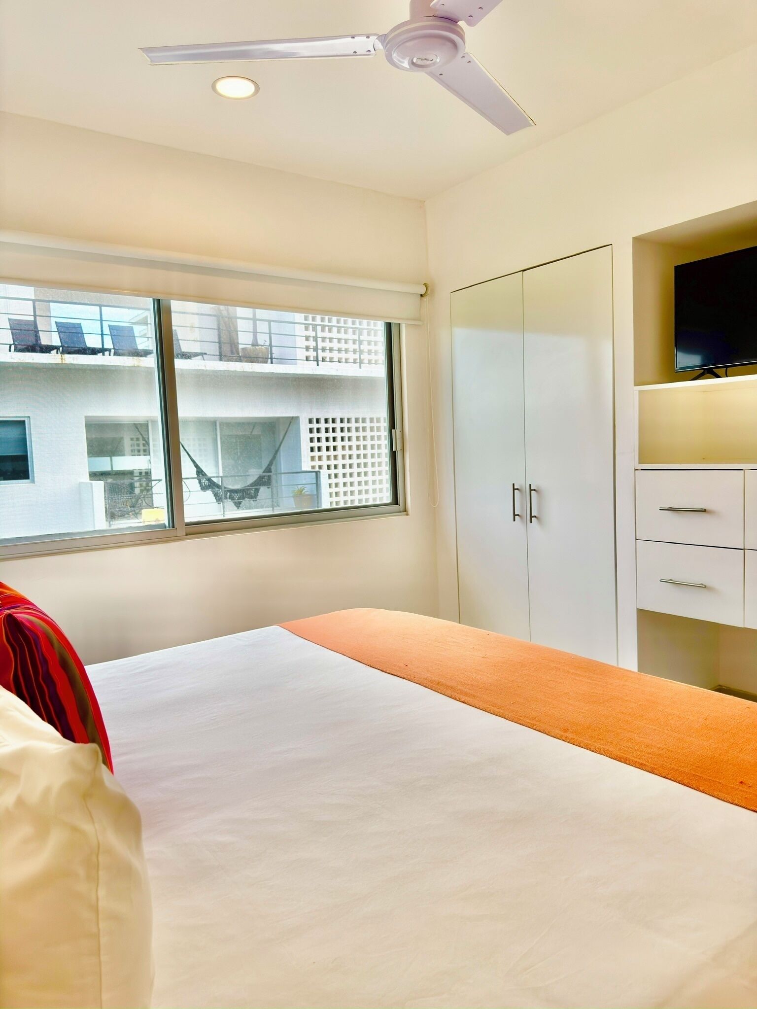 A bedroom with a bed, a ceiling fan, a TV, and a window with a view of buildings.