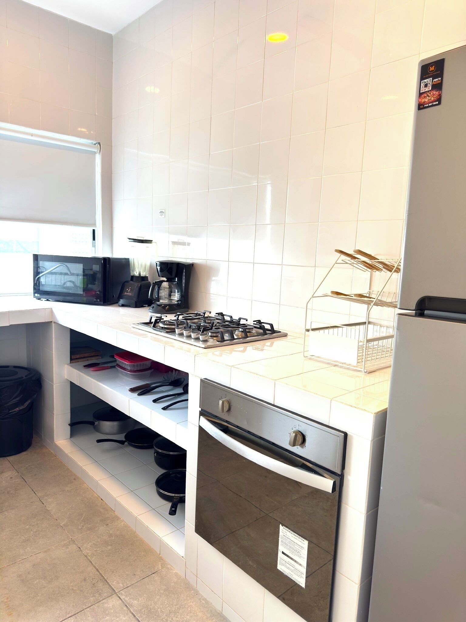 A compact kitchen with white tiled walls, a gas stove, a microwave, a coffee maker, and a refrigerator.