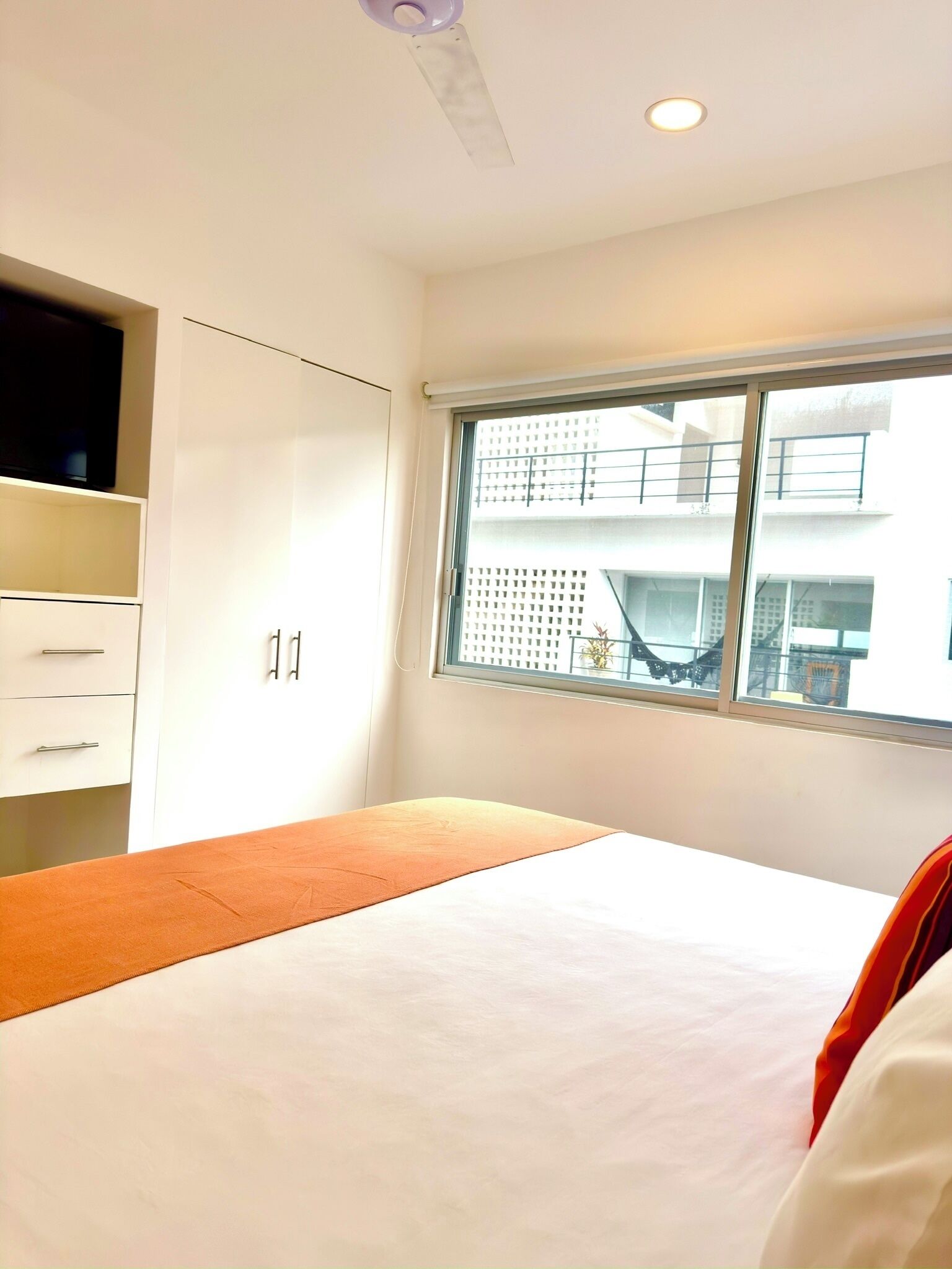 Un dormitorio con cama, ventana con vista a edificios y ventilador de techo.