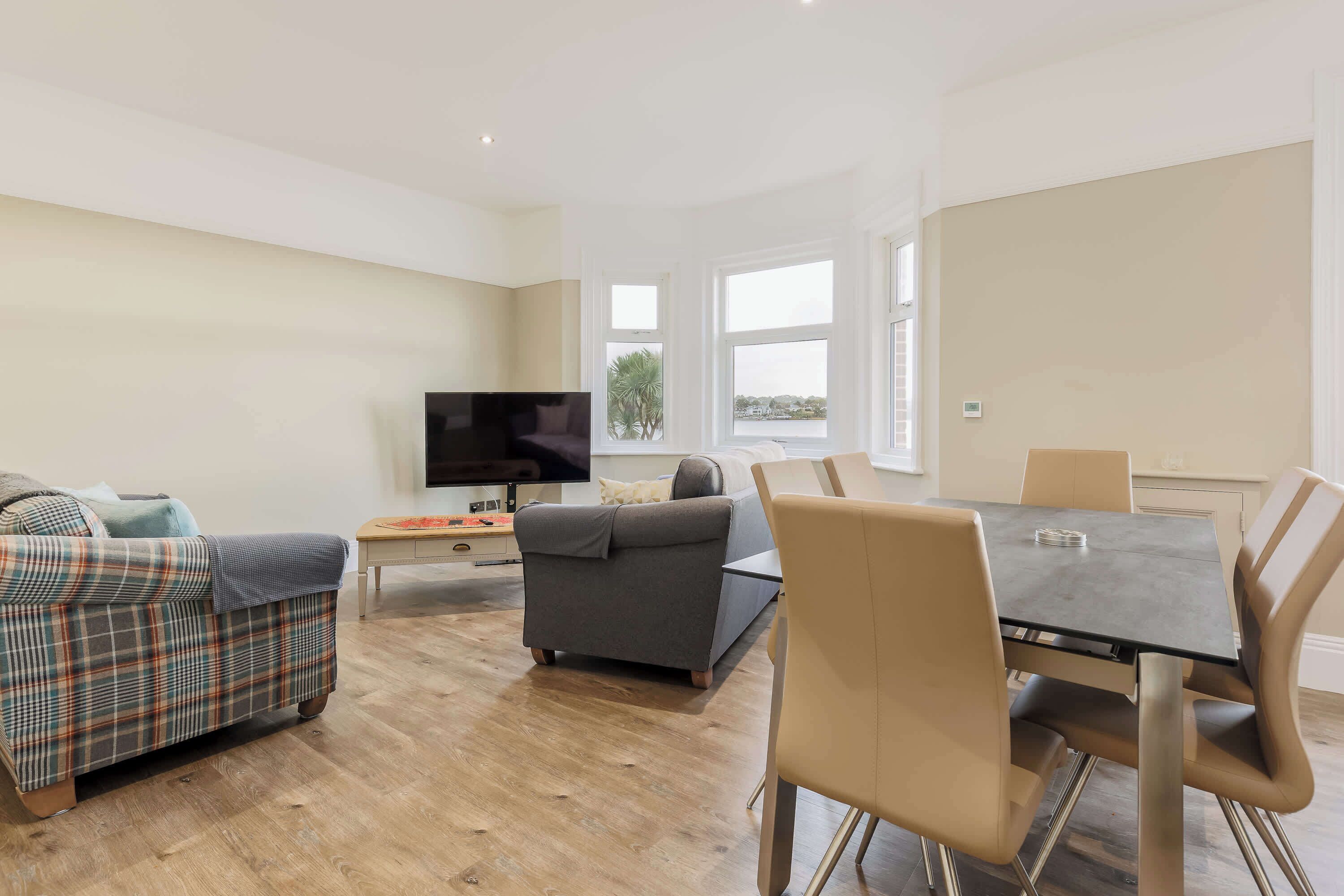 A modern living room with a sofa, a dining table with chairs, a TV, and large windows with a view.