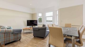 A modern living room with a sofa, a dining table with chairs, a TV, and large windows with a view.