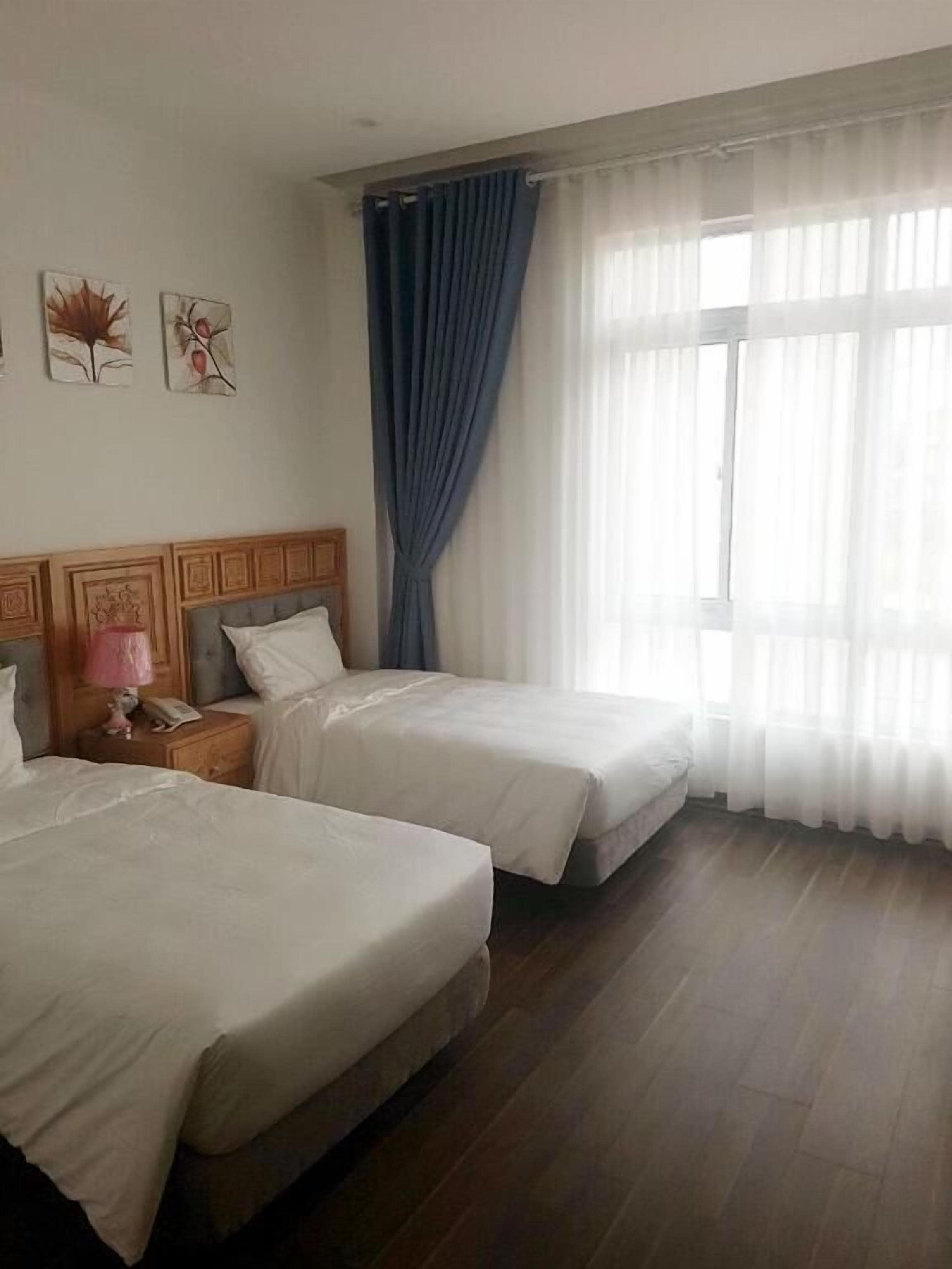 Una habitación de hotel con dos camas, un cabecero de madera, una mesita de noche con una lámpara y una ventana con cortinas traslúcidas.