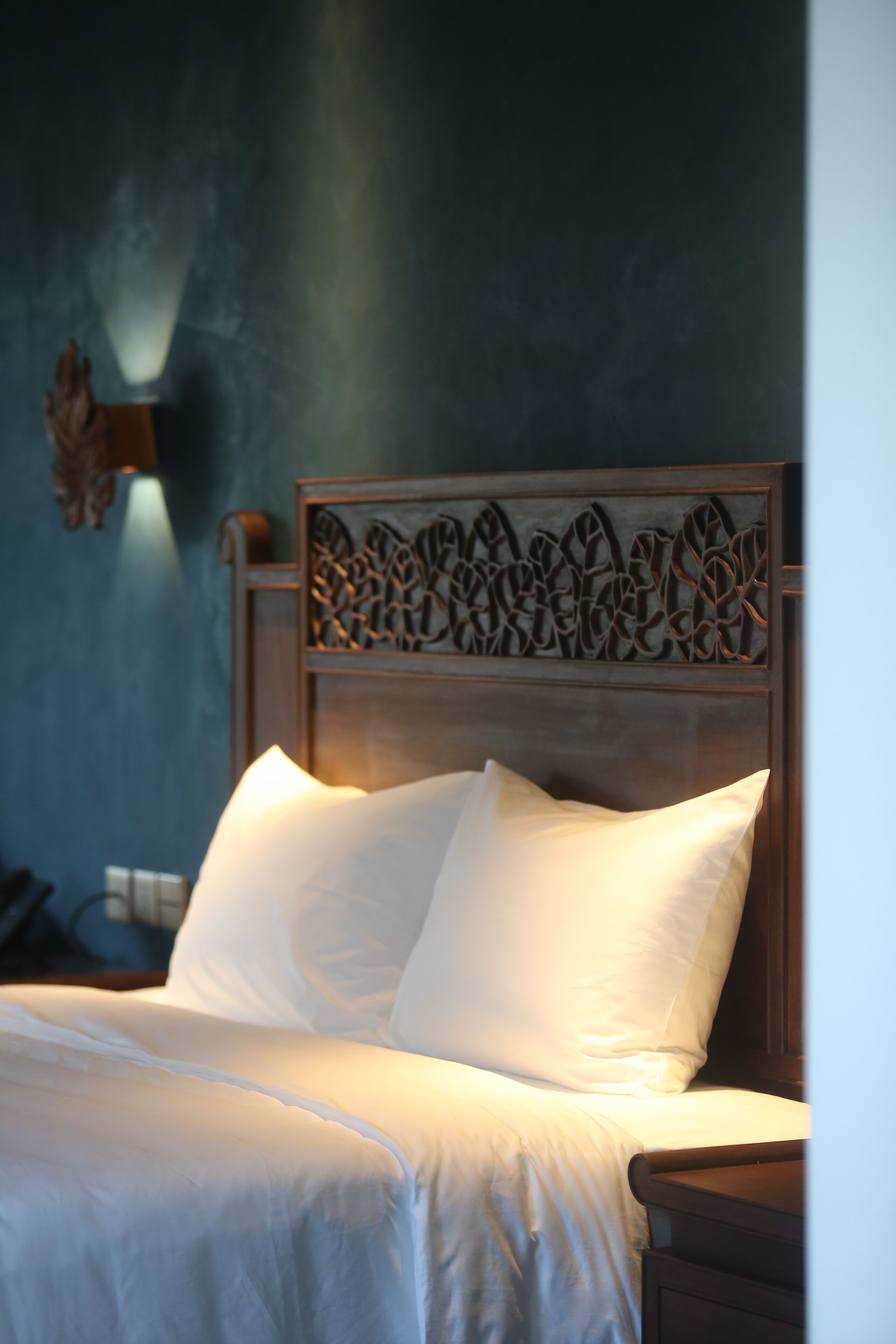 Uma cama bem arrumada com lençóis e almofadas brancas, um painel decorativo e uma luminária fixada na parede.