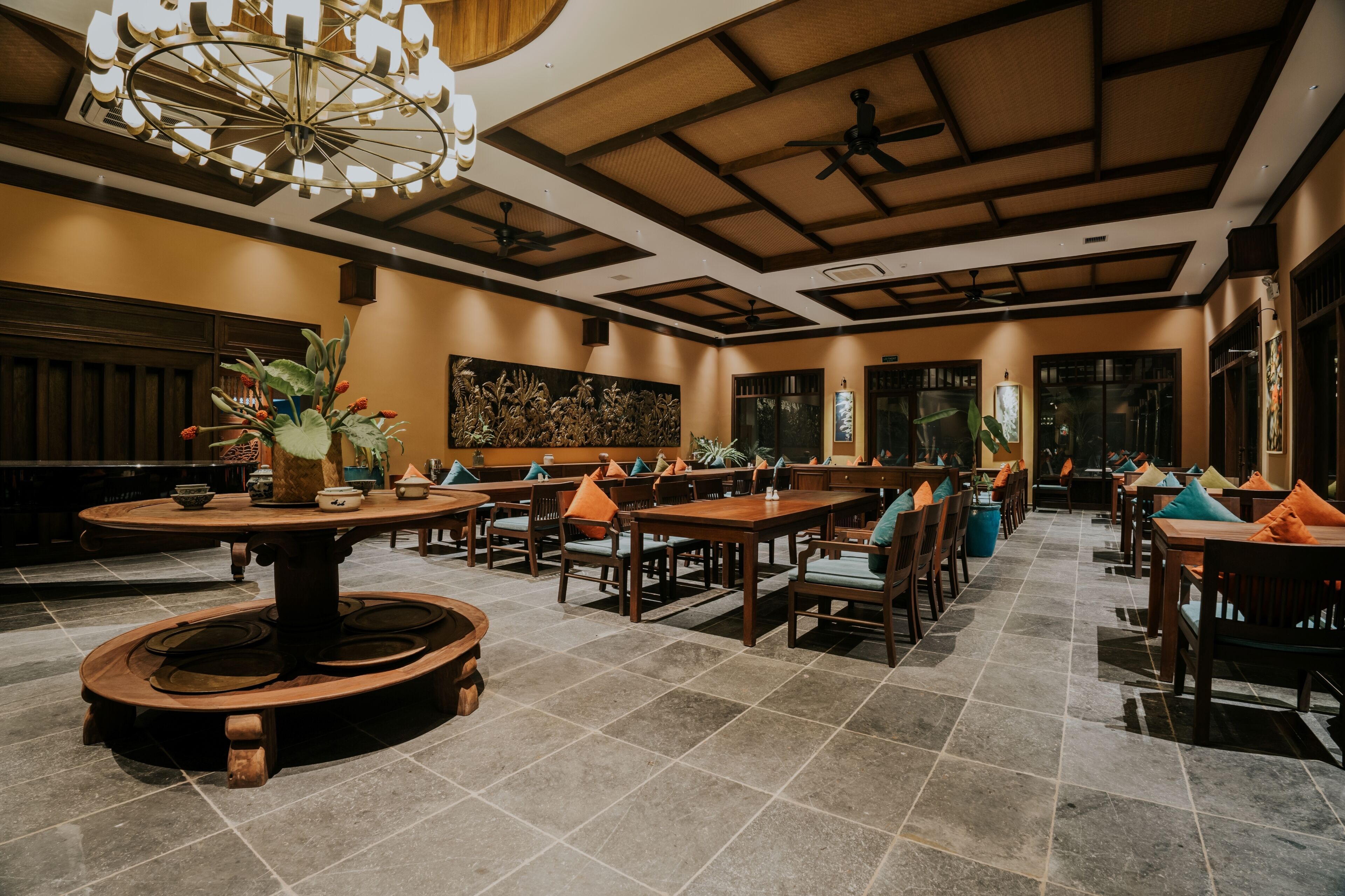 A spacious restaurant with high ceilings, wooden tables and chairs, and a central round table with a flower arrangement.