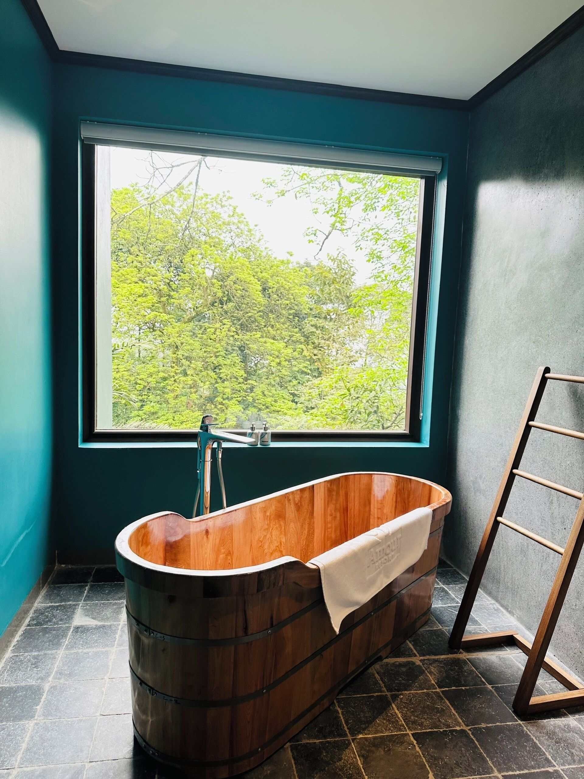A wooden bathtub in a room with teal walls and a large window overlooking a forest.