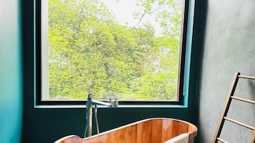 A wooden bathtub in a room with teal walls and a large window overlooking a forest.