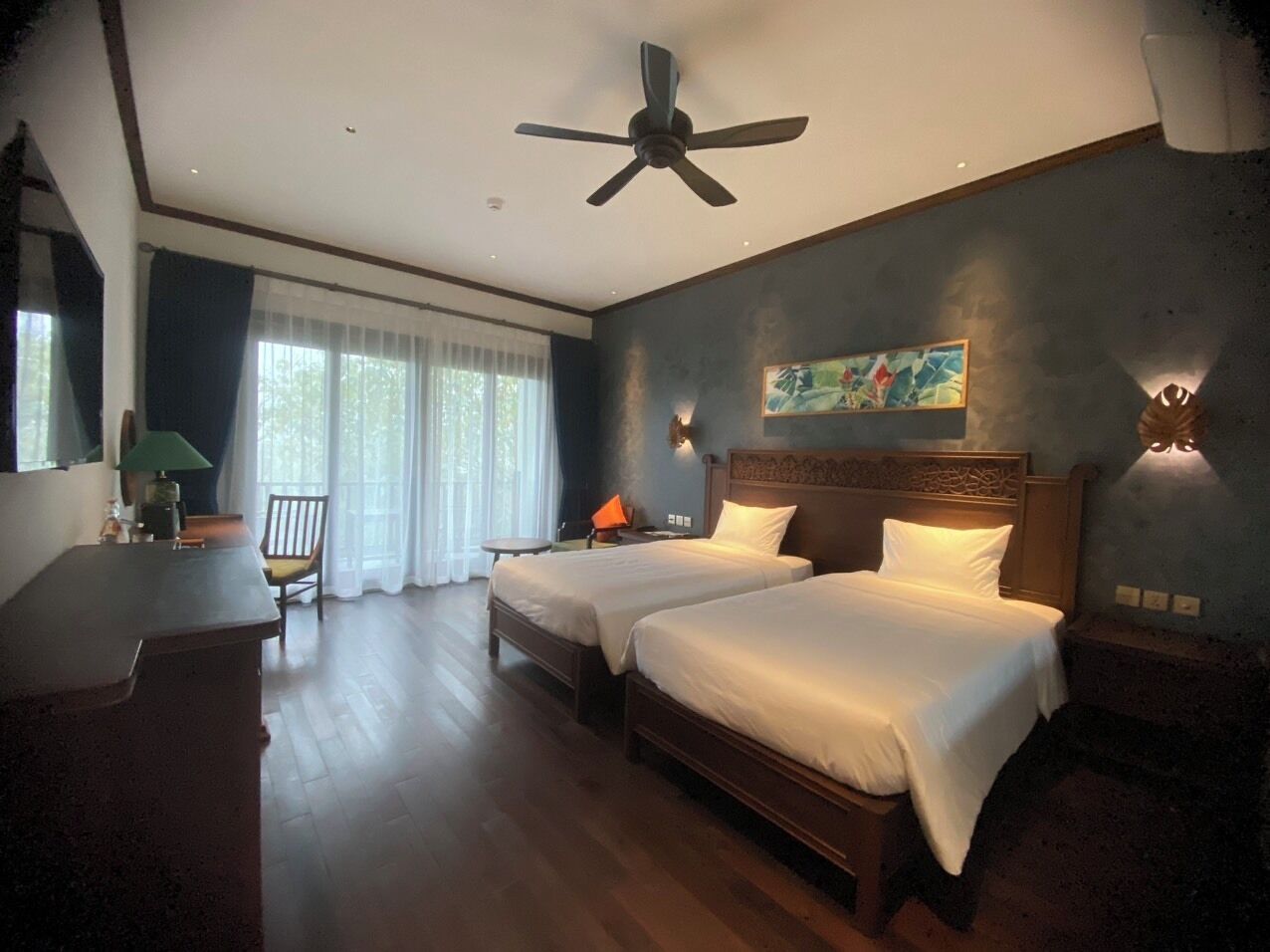 A hotel room with two beds, a desk, a chair, a ceiling fan, and a painting on the wall.