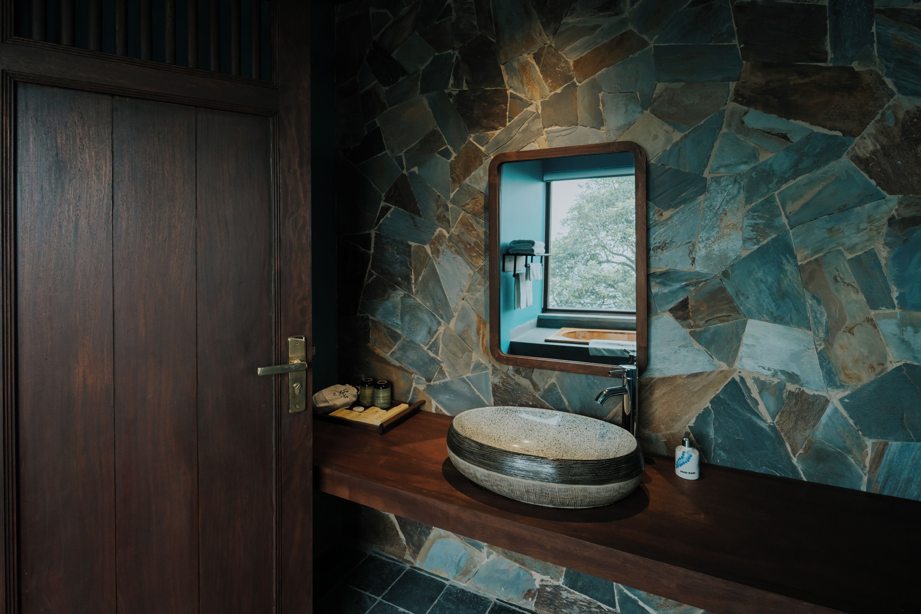 A bathroom with a stone wall, a wooden door, and a round sink with a mirror above it.