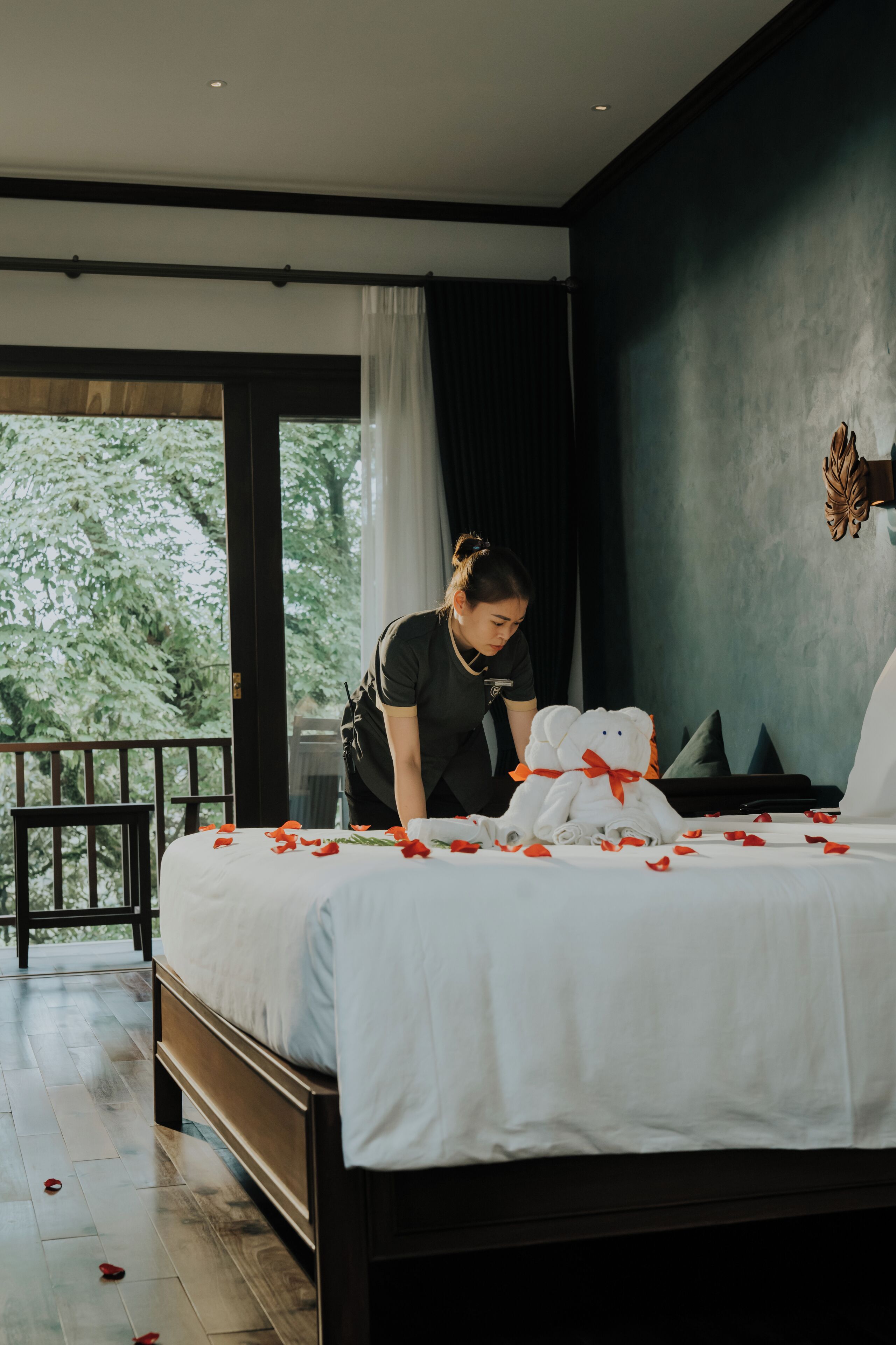 A hotel room with a bed, a large stuffed teddy bear, and a view of trees through a window.