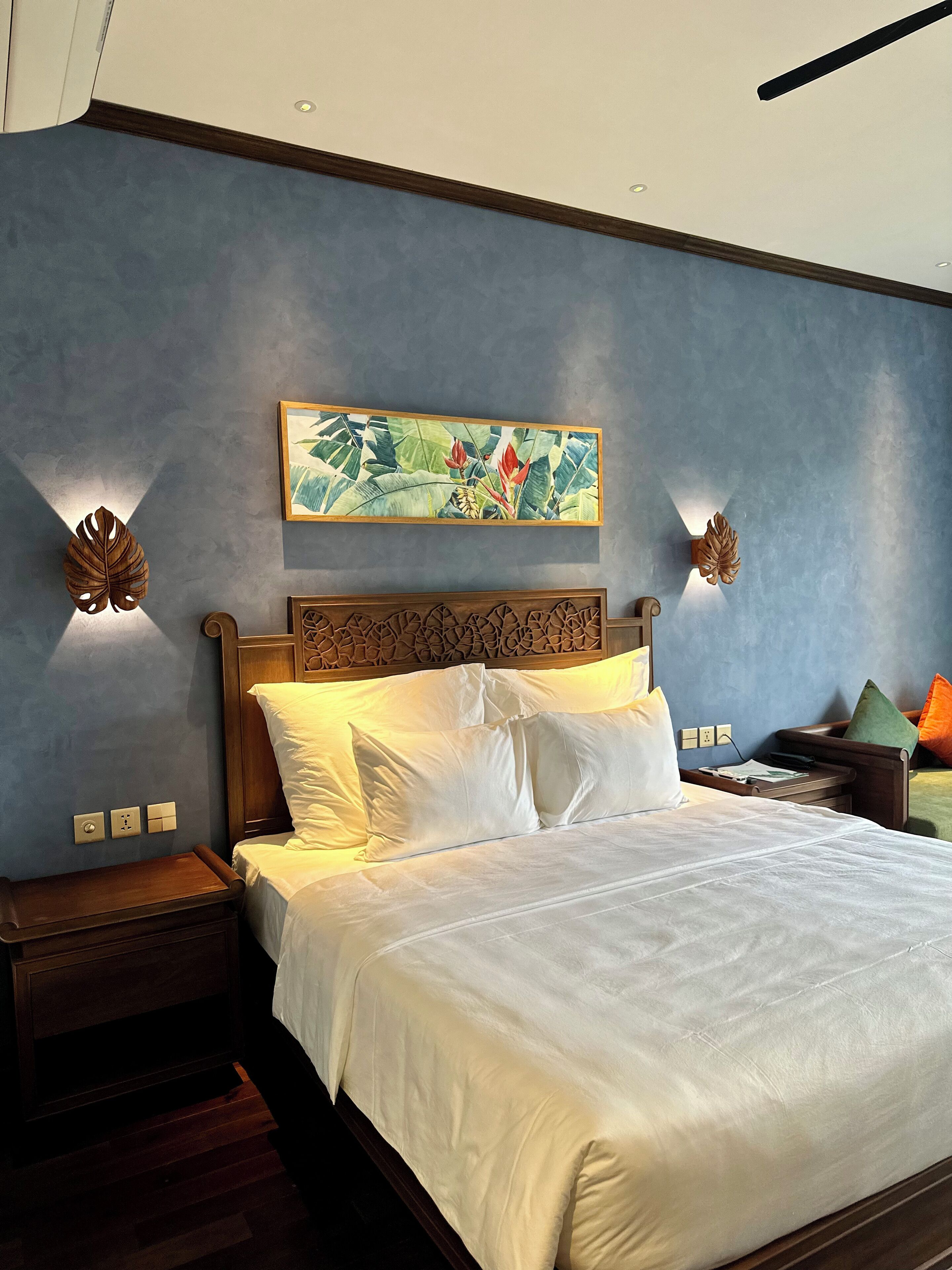 A neatly made bed with white linens, a wooden headboard, and a tropical-themed painting above it.
