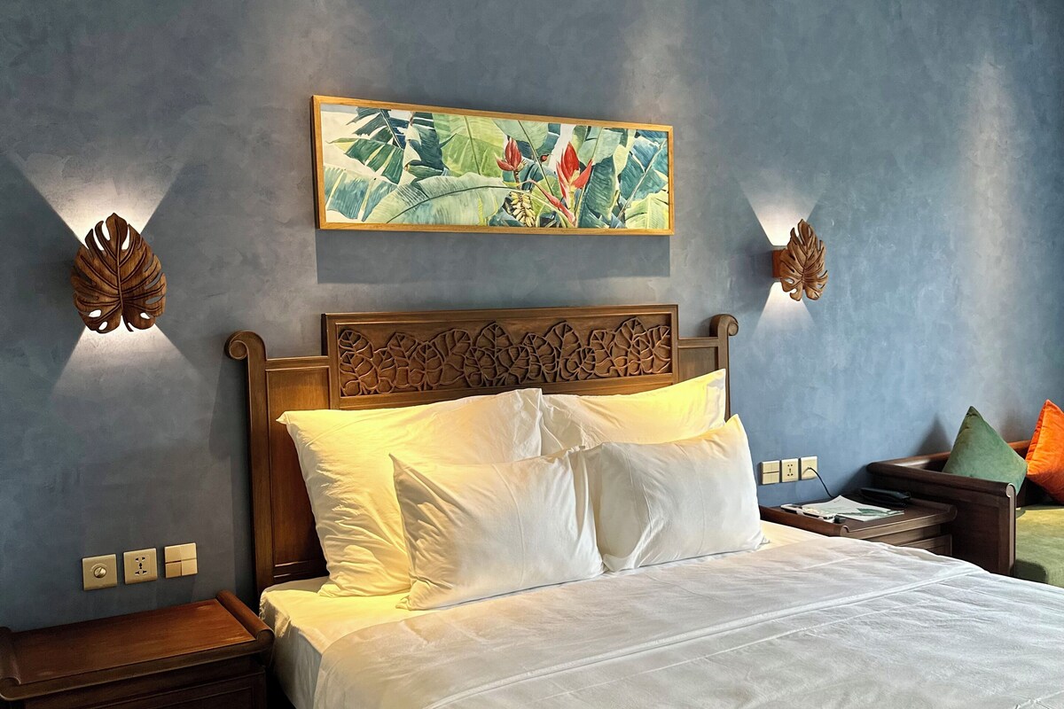 A neatly made bed with white linens, a wooden headboard, and a tropical-themed painting above it.