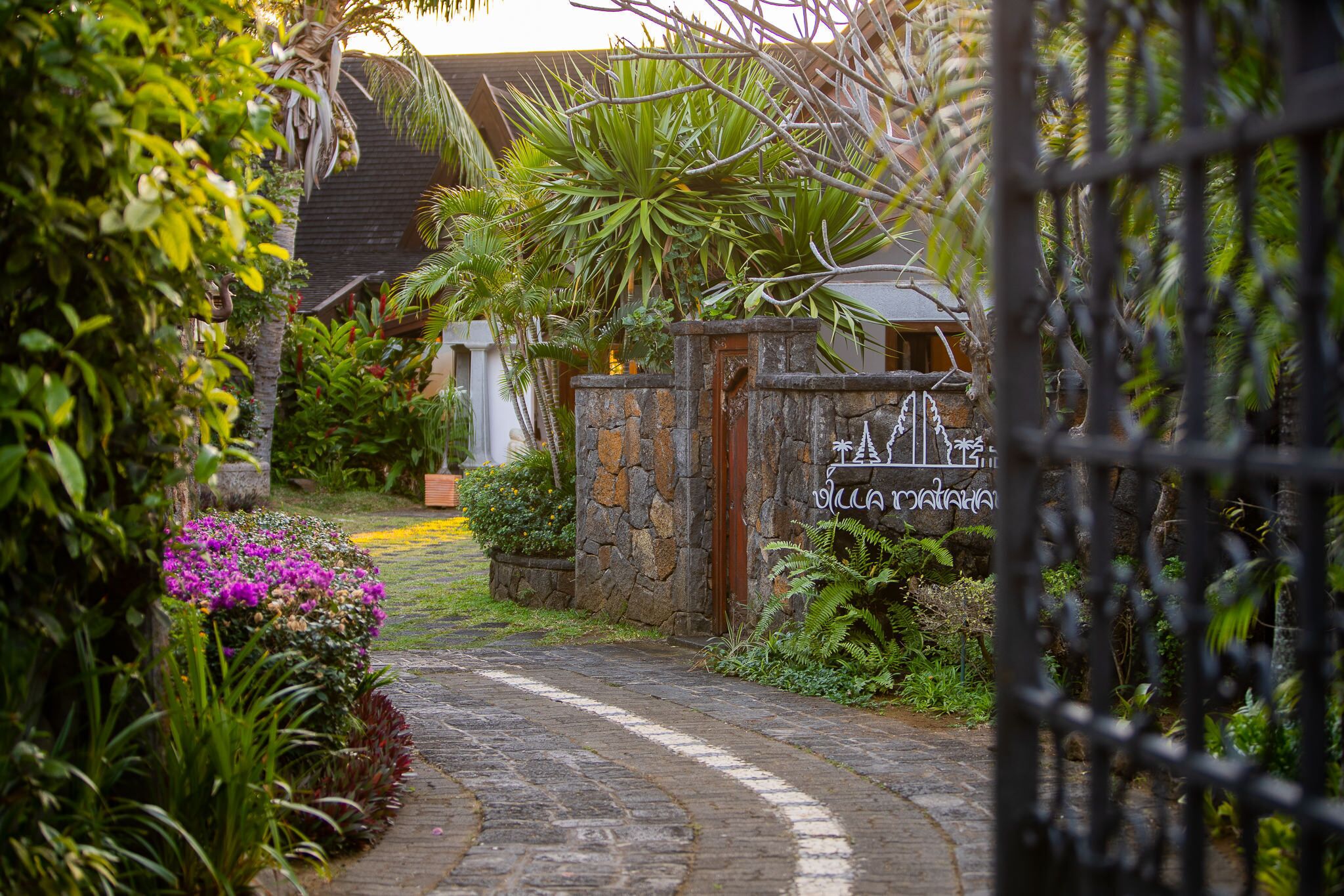A stone gate with a sign, surrounded by lush greenery and a stone pathway leading to a building.