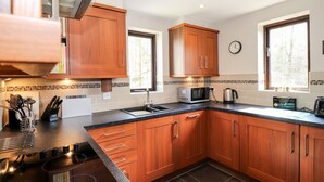 A modern kitchen with wooden cabinets, a microwave, a kettle, and a window with a view of trees. - Ghyll View (Ambleside)