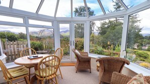 Ein Wintergarten mit Tisch und Stühlen, einem Korbsessel und Blick auf Garten und Berge.