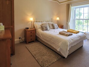 A neatly arranged bedroom with a large bed, two bedside tables with lamps, a wooden nightstand, a beige rug, and a window with curtains. - Sandburne Cottage (Keswick)