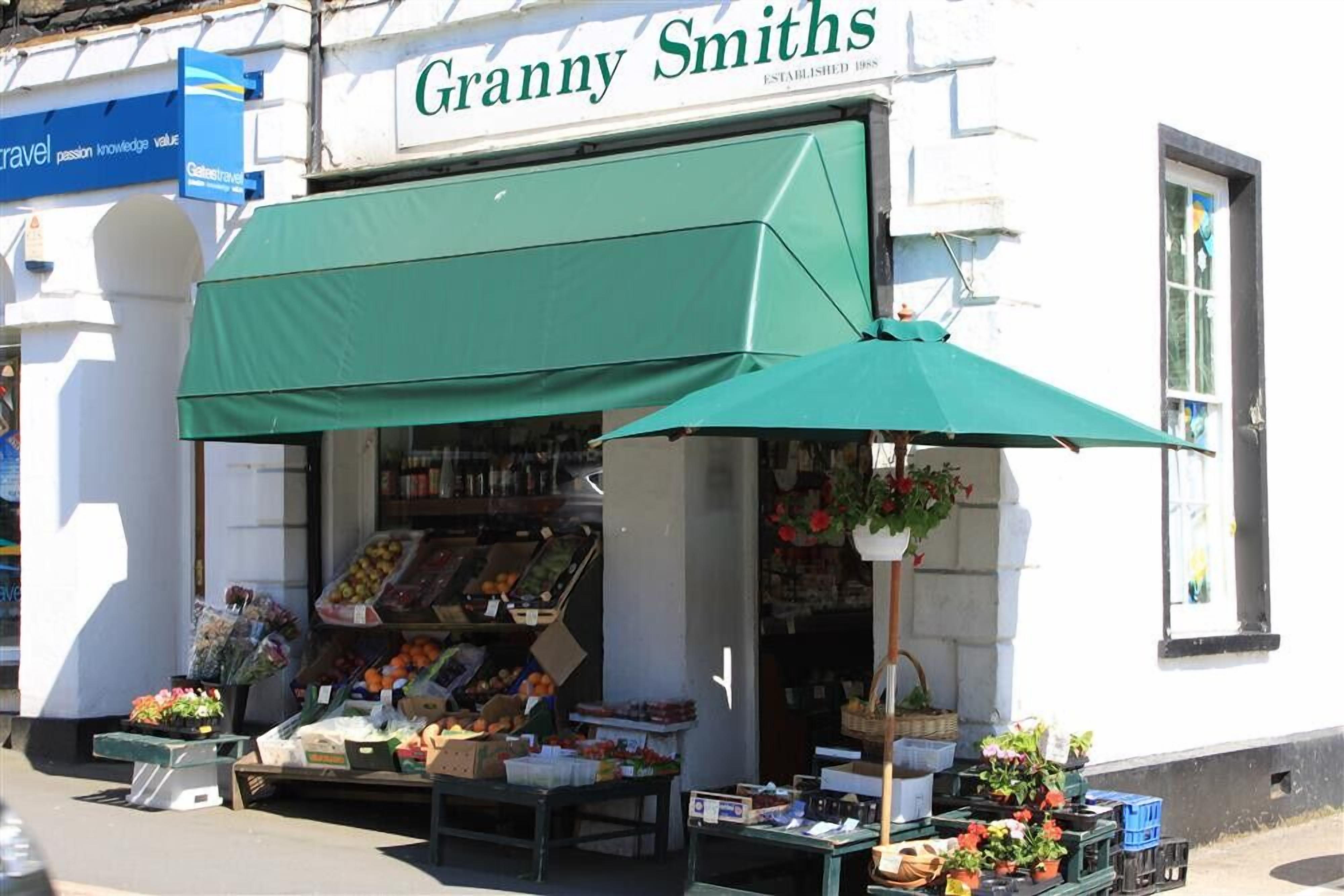Une devanture de magasin avec un auvent vert, une variété de fruits et de légumes exposés, et une corbeille de fleurs suspendue.