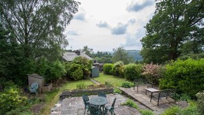Une terrasse en pierre avec une table ronde et des chaises, entourée d’une végétation luxuriante et offrant une vue sur un paysage vallonné.