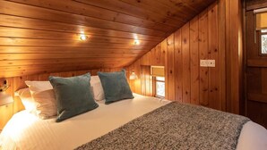 Ein gemütliches Schlafzimmer mit Holzwänden, einem Bett mit weißer Bettwäsche und zwei türkisfarbenen Kissen, einer grauen Tagesdecke und einem Fenster mit Blick.