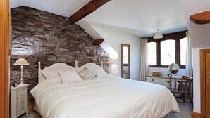 Un dormitorio con pared de piedra, una cama con ropa de cama blanca, una mesita de noche con lámpara, un escritorio de madera con espejo y una ventana con cortinas.