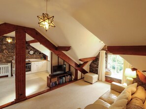 A loft-style living room with exposed wooden beams, a stone wall, a fireplace, a television, and a cozy seating area. - High Raise (Ambleside)