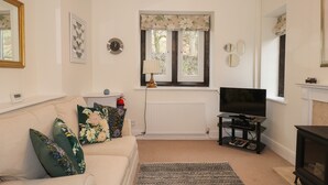 A living room with a sofa, a television, a fireplace, and a window with a floral valance. - Larch Cottage (Ambleside)
