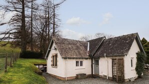 A white house with a gravel driveway, a wooden fence, and a large tree in the background. - Larch Cottage (Ambleside)